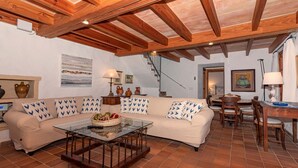 A cozy living room with a wooden ceiling, a sofa with patterned cushions, a glass coffee table, and a dining area with wooden furniture. - Finca Gloria for 4 persons with pool (Pollensa)
