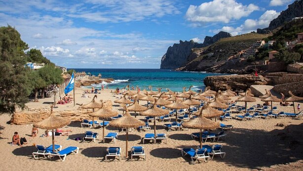A beach with sun loungers and thatched umbrellas, rocky cliffs, and buildings in the background. - Finca Gloria for 4 persons with pool (Pollensa)