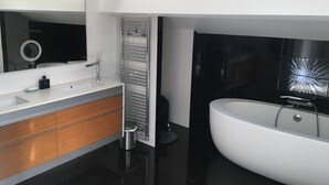A modern bathroom with a large white bathtub, a built-in sink, and a vanity with wooden drawers. - House the Horsemen (Anglet)