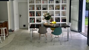 A modern living room with a large white bookshelf filled with books, a dining table with chairs, and a large glass door leading to an outdoor area. - House the Horsemen (Anglet)