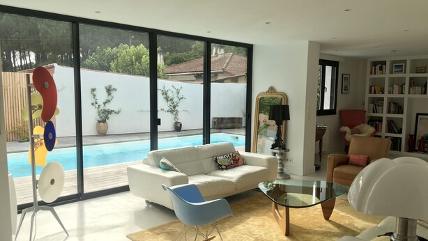 A modern living room with a white sofa, a blue rocking chair, a glass coffee table, and a large window overlooking a swimming pool. - House the Horsemen (Anglet)