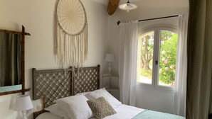 A bedroom with a bed, a nightstand, a lamp, a dreamcatcher, and a window with curtains. - Le Mas Mellou (Orange)