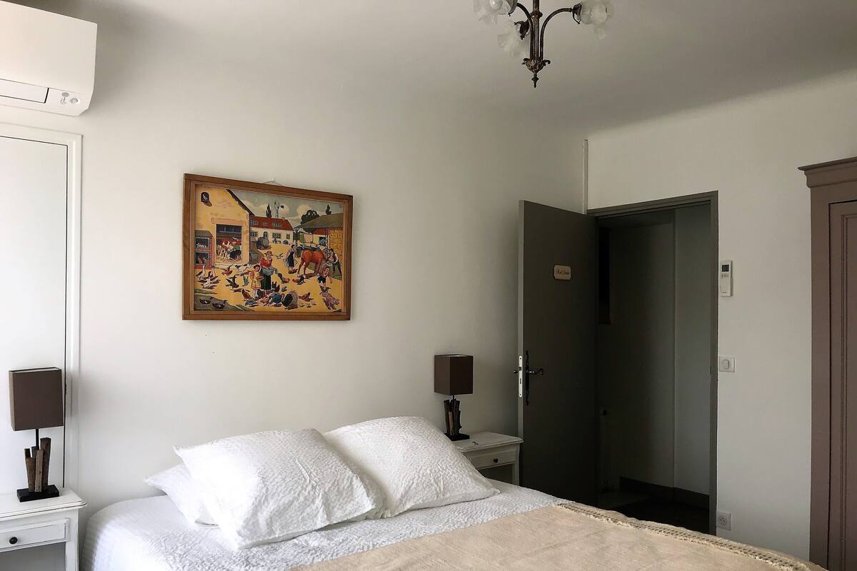 A bedroom with a bed, a bedside table, a lamp, a painting, and a door.