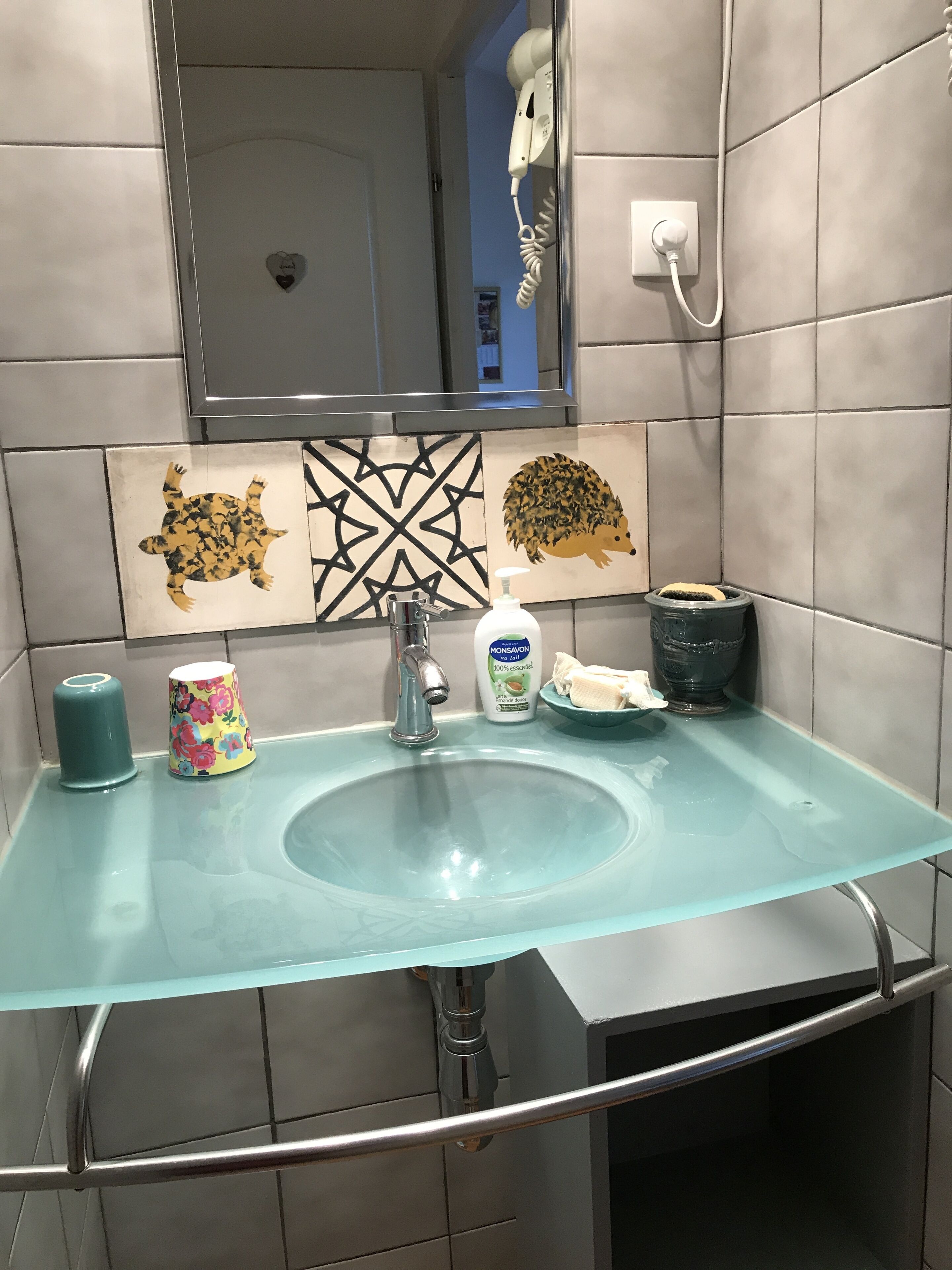 Une salle de bain avec un lavabo en verre, un miroir, un sèche-cheveux et des carreaux décoratifs.