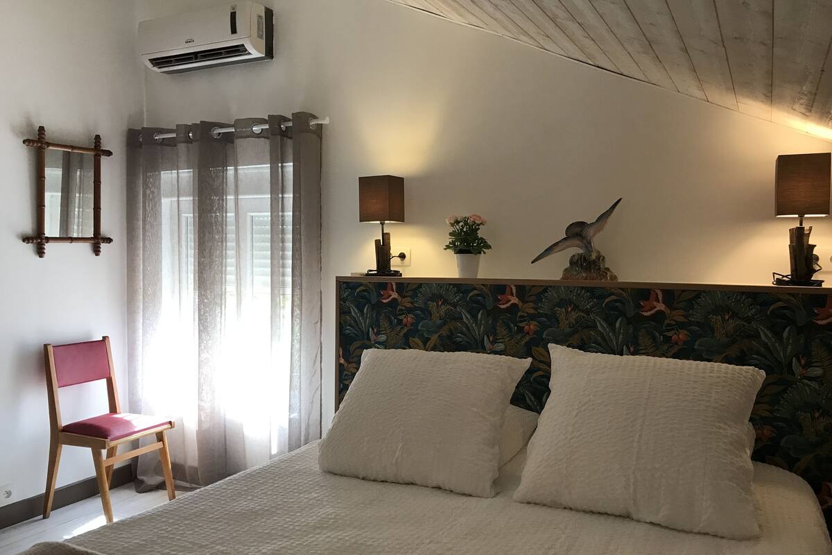 A bedroom with a bed, two bedside lamps, a wall-mounted air conditioner, a wooden chair, and a window with curtains.
