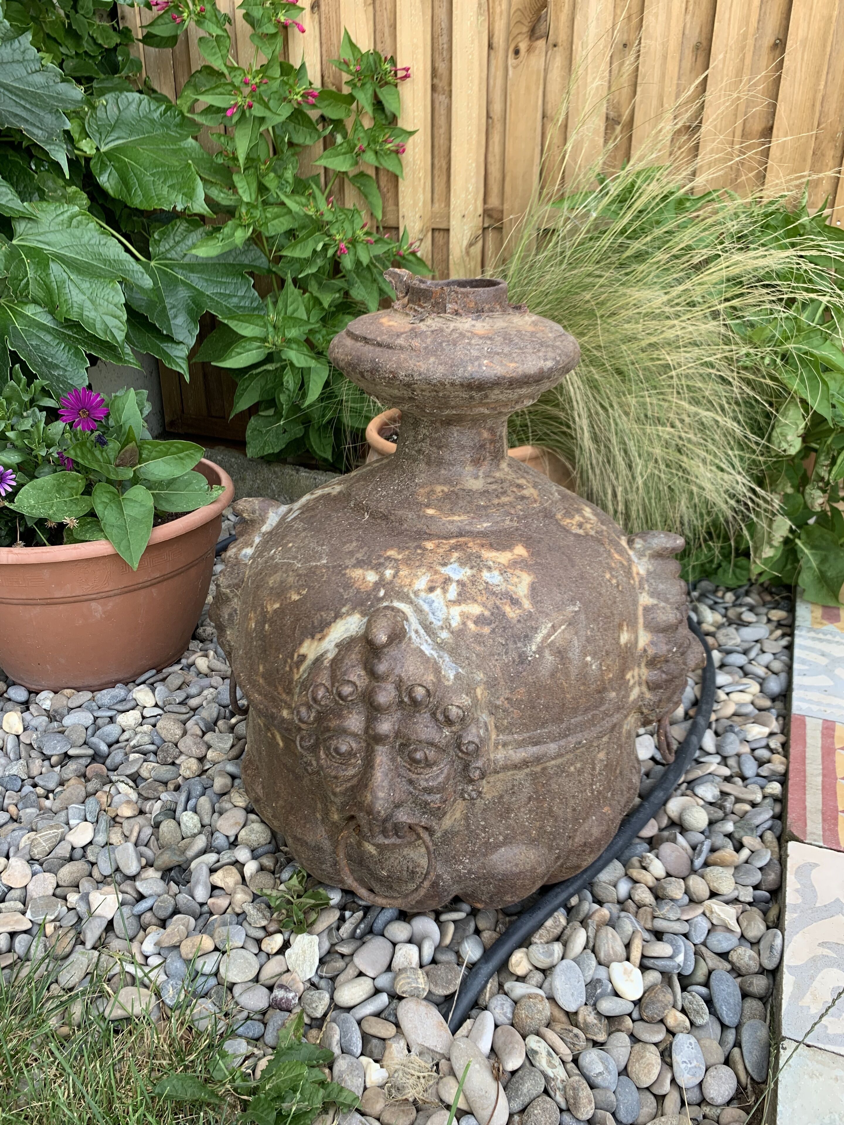 A large, weathered stone urn with a lid, surrounded by pebbles and greenery.