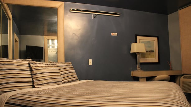 A hotel room with a bed, a bedside table, a lamp, and a wall-mounted shelf. - Royal Budget Inn (Redford)