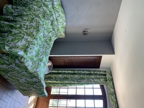 Une chambre avec un lit recouvert d’une literie à motifs de feuilles vertes, un ventilateur de plafond, une porte en bois et une fenêtre donnant sur un paysage verdoyant.
