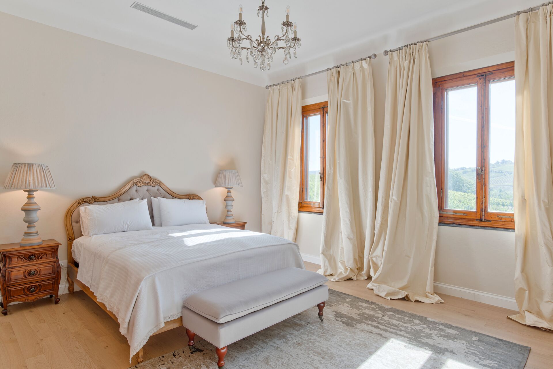 Ein Schlafzimmer mit einem großen Bett, zwei Nachttischen mit Lampen, einer Bank, einer Chandelier und einem Fenster mit Vorhängen.