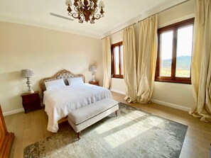 A bedroom with a large bed, a chandelier, a nightstand, and a view of the outdoors through a window.