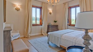 A bedroom with a bed, a dresser, a bench, a chandelier, and two windows with curtains.