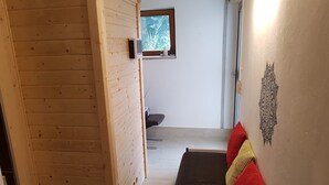 A room with a wooden floor, a black sofa with red and yellow cushions, a small window, and a wall with a decorative pattern. - With us (Tittling)