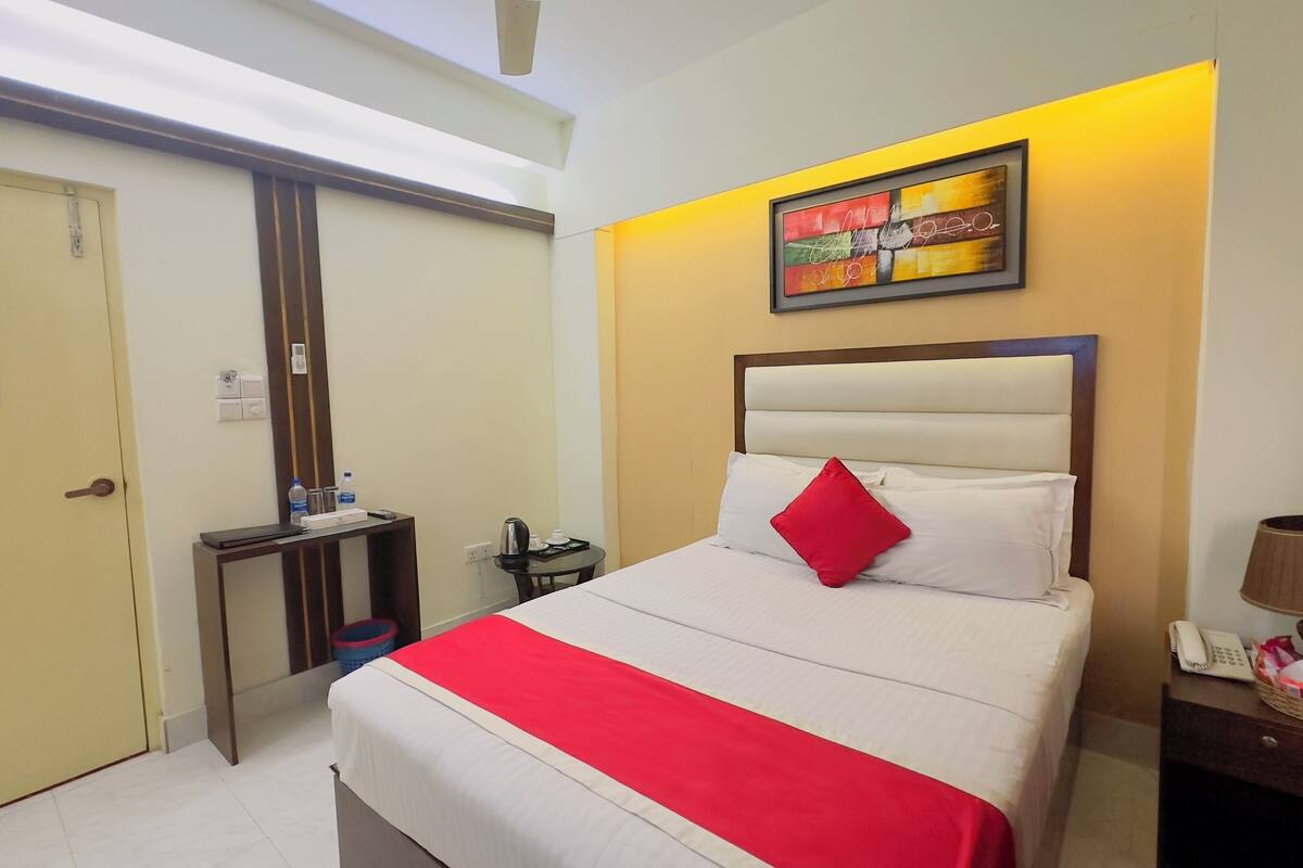 A hotel room with a bed, a bedside table with a lamp, a small table with a kettle, and a wall-mounted artwork.