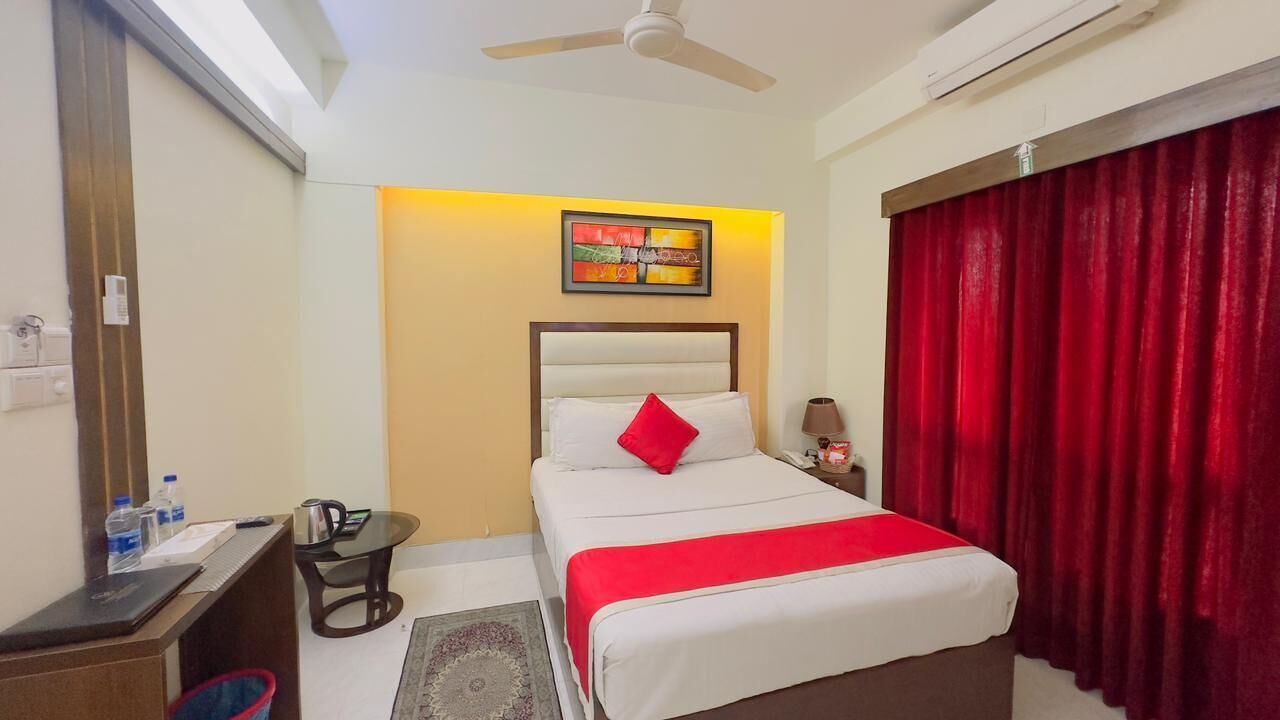 A hotel room with a bed, a desk, a chair, a ceiling fan, and a red curtain.