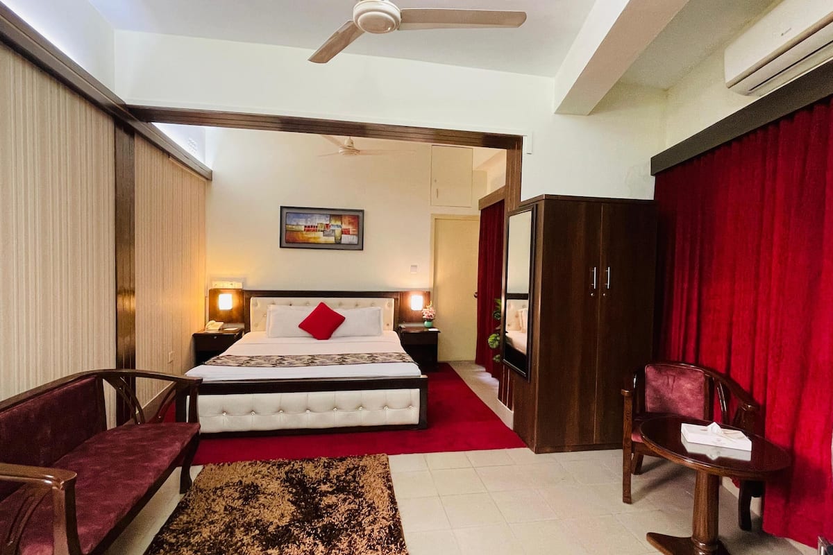 A hotel room with a bed, two chairs, a red curtain, a painting on the wall, and a ceiling fan.