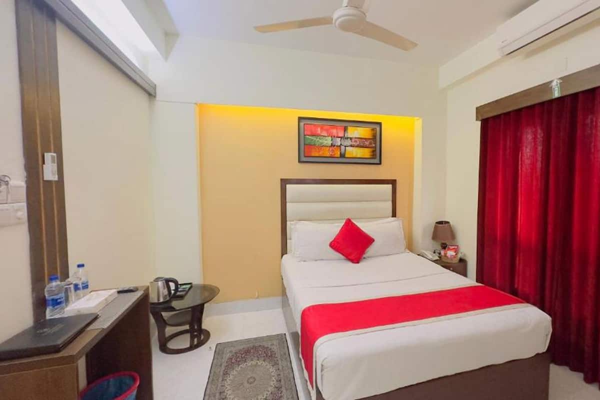 A hotel room with a bed, a desk, a chair, a nightstand, a red curtain, a ceiling fan, and a painting on the wall.