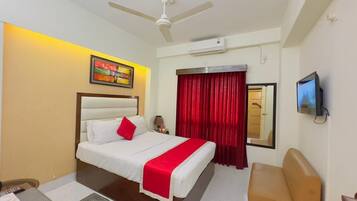 A hotel room with a bed, a desk, a chair, a ceiling fan, a TV, and a window with red curtains.