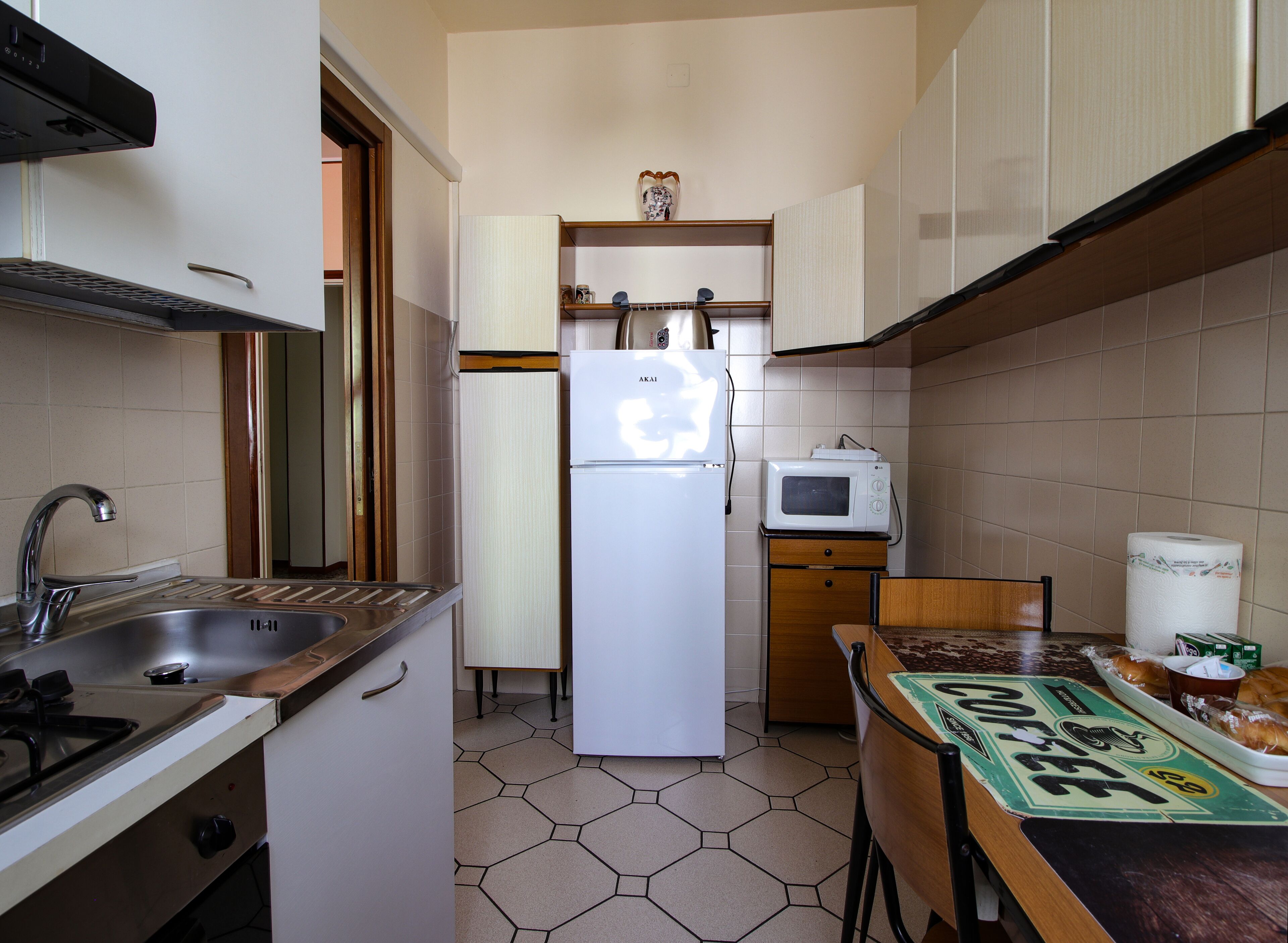 Una cocina compacta con lavamanos, estufa, refrigerador, microondas y una pequeña mesa de madera con un mantel individual de tema de café.