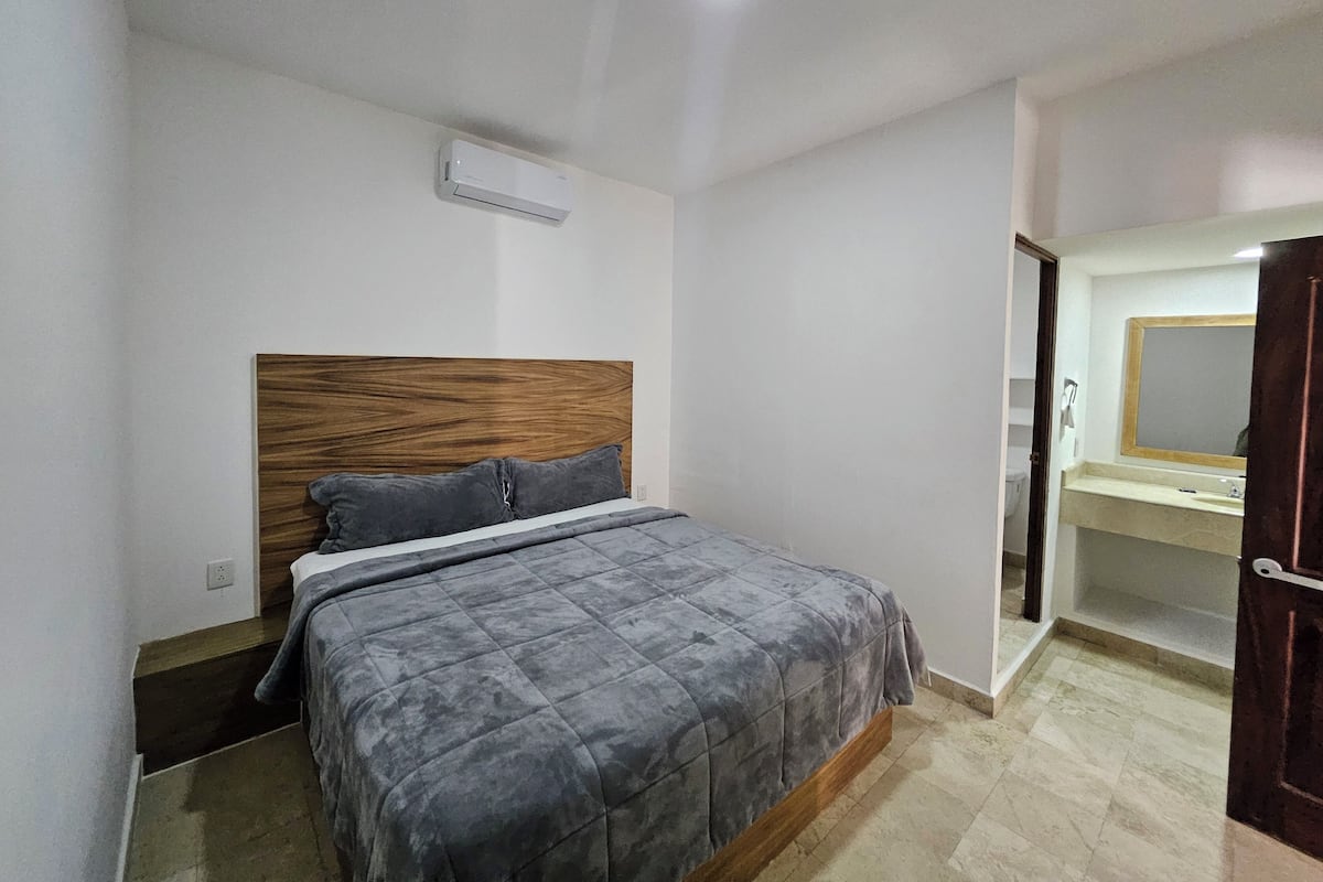 Un dormitorio con cama, cabecera de madera, colcha acolchada gris, pared blanca, luz de techo y baño con espejo y lavabo.