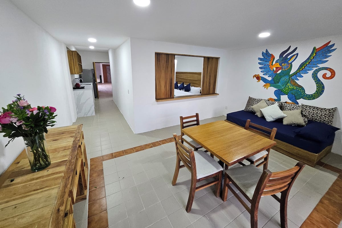 Un comedor con una mesa y sillas de madera, un sofá con cojines estampados y un mural decorativo en la pared.