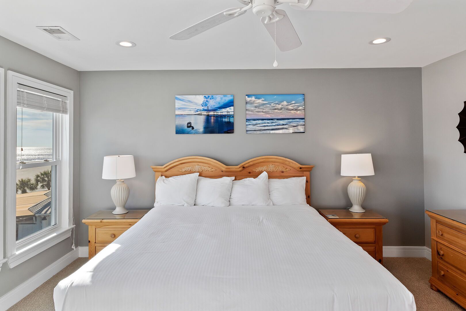 A bedroom with a large bed, two bedside tables with lamps, a ceiling fan, and two wall-mounted pictures of a beach scene.