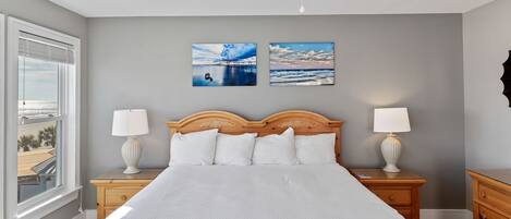 A bedroom with a large bed, two bedside tables with lamps, a ceiling fan, and two wall-mounted pictures of a beach scene.