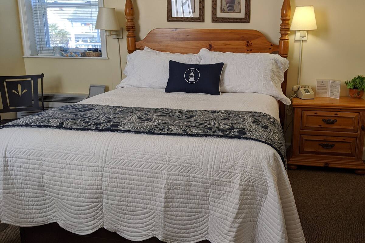 Un lit bien fait, recouvert d’une couette blanche et agrémenté d’un coussin décoratif, une table de chevet en bois avec une lampe, et une petite plante en pot.