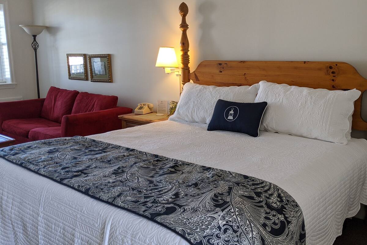 Un lit bien fait, avec une couvre-lit à motifs, une tête de lit en bois et un fauteuil rouge, dans une chambre d’hôtel.