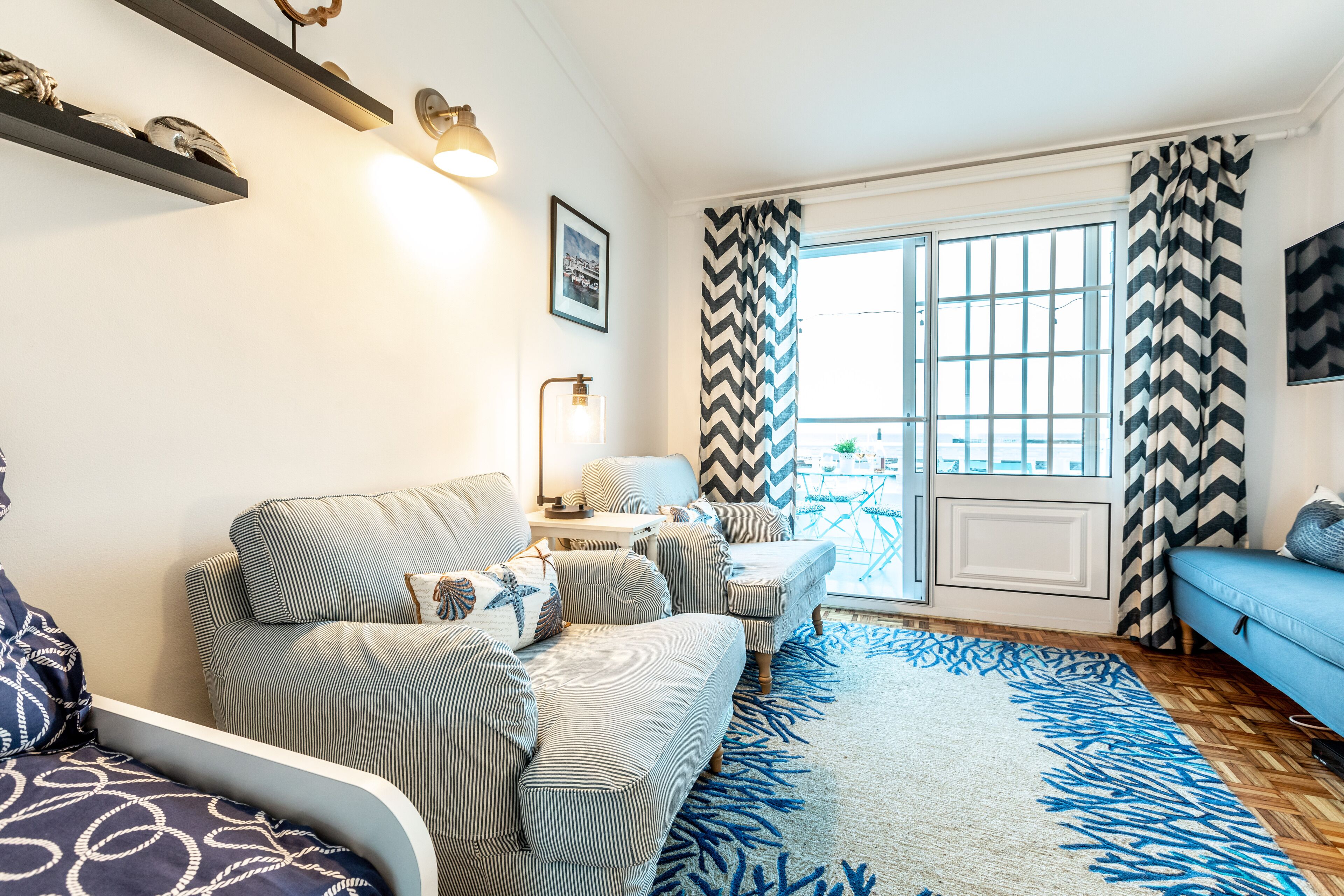 Ein Wohnzimmer mit Sofa, Sesseln, Couchtisch und Fernseher. Ein Fenster mit Vorhängen und Ausblick nach draußen.