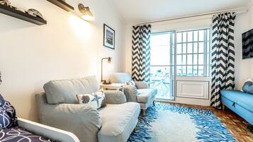 Ein Wohnzimmer mit Sofa, Sesseln, Couchtisch und Fernseher. Ein Fenster mit Vorhängen und Ausblick nach draußen.