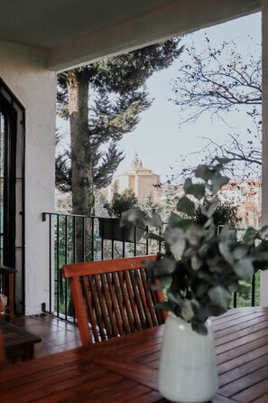 A wooden table with a white vase of green plants, a wooden bench, and a view of a tree and buildings in the background.