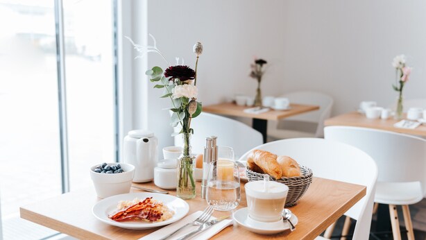 A breakfast setting with a wooden table, white chairs, a bowl of blueberries, a plate of bacon, a croissant, a vase with flowers, a candle, and a glass of milk. - HAIMHAUSERS Hotel Garni (Haimhausen)