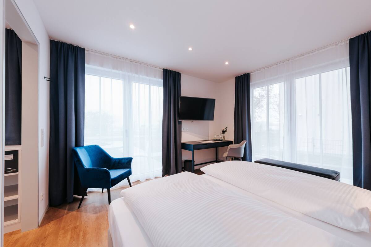 A hotel room with two beds, a desk with a chair, a TV, and large windows with curtains.
