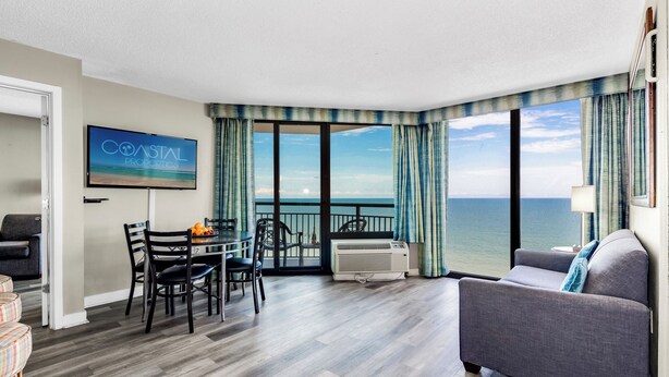 A hotel room with a dining area, a sofa, a TV displaying 'COASTAL PROPERTIES', and a view of the ocean. - 17th Floor 1Br Oceanfront/ Dogs OK! (Myrtle Beach)