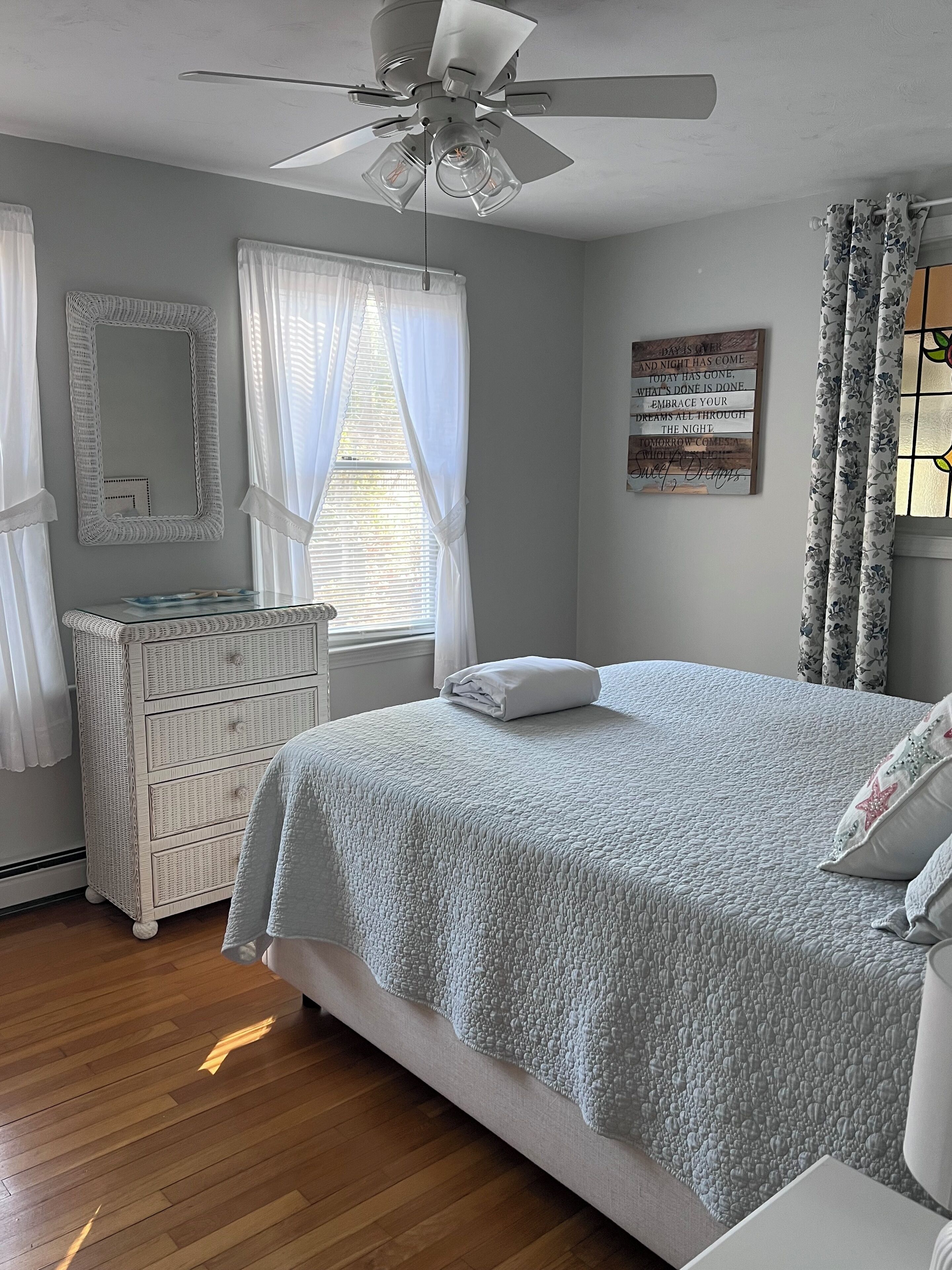 Un dormitorio con cama, cómoda, ventilador de techo y ventana con cortinas.