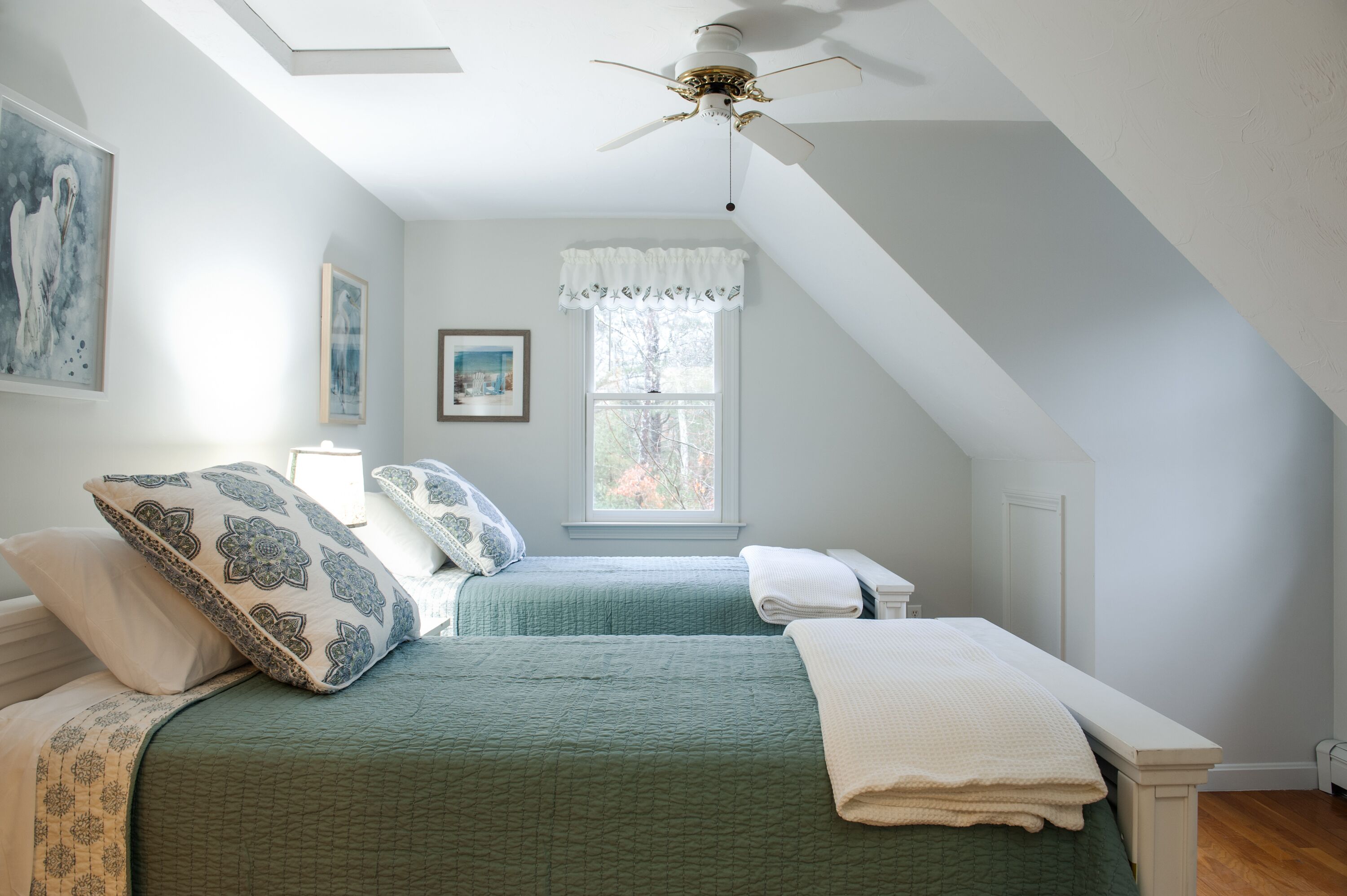 Ein Schlafzimmer mit zwei Betten, einem Deckenventilator, einem Fenster mit weißen Vorhängen und Bildern an den Wänden.