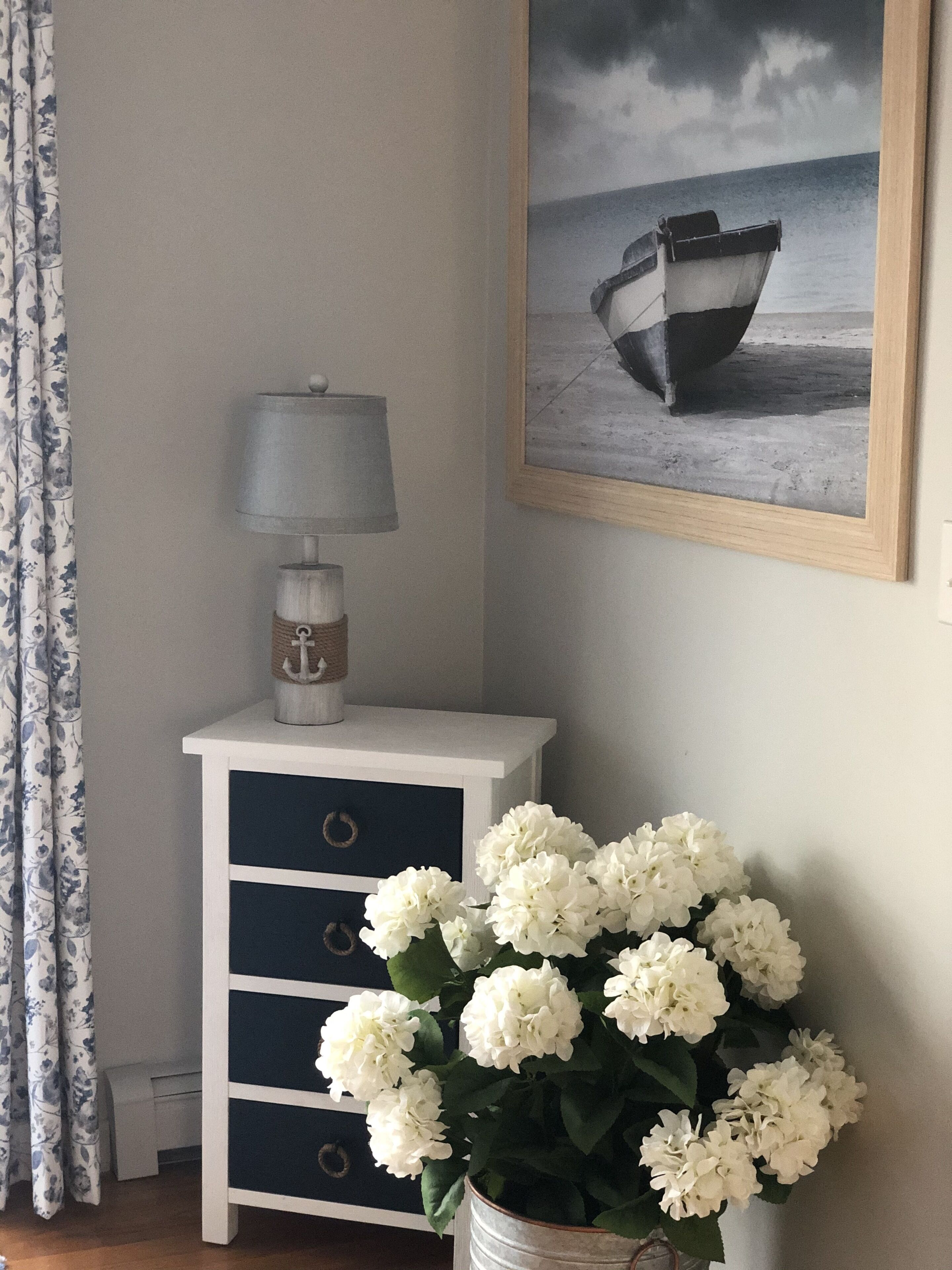 Et hjørne af et rum med et hvidt natbord, en lampe, et billedramme med et skib på vandet, og en vase med hvide blomster.