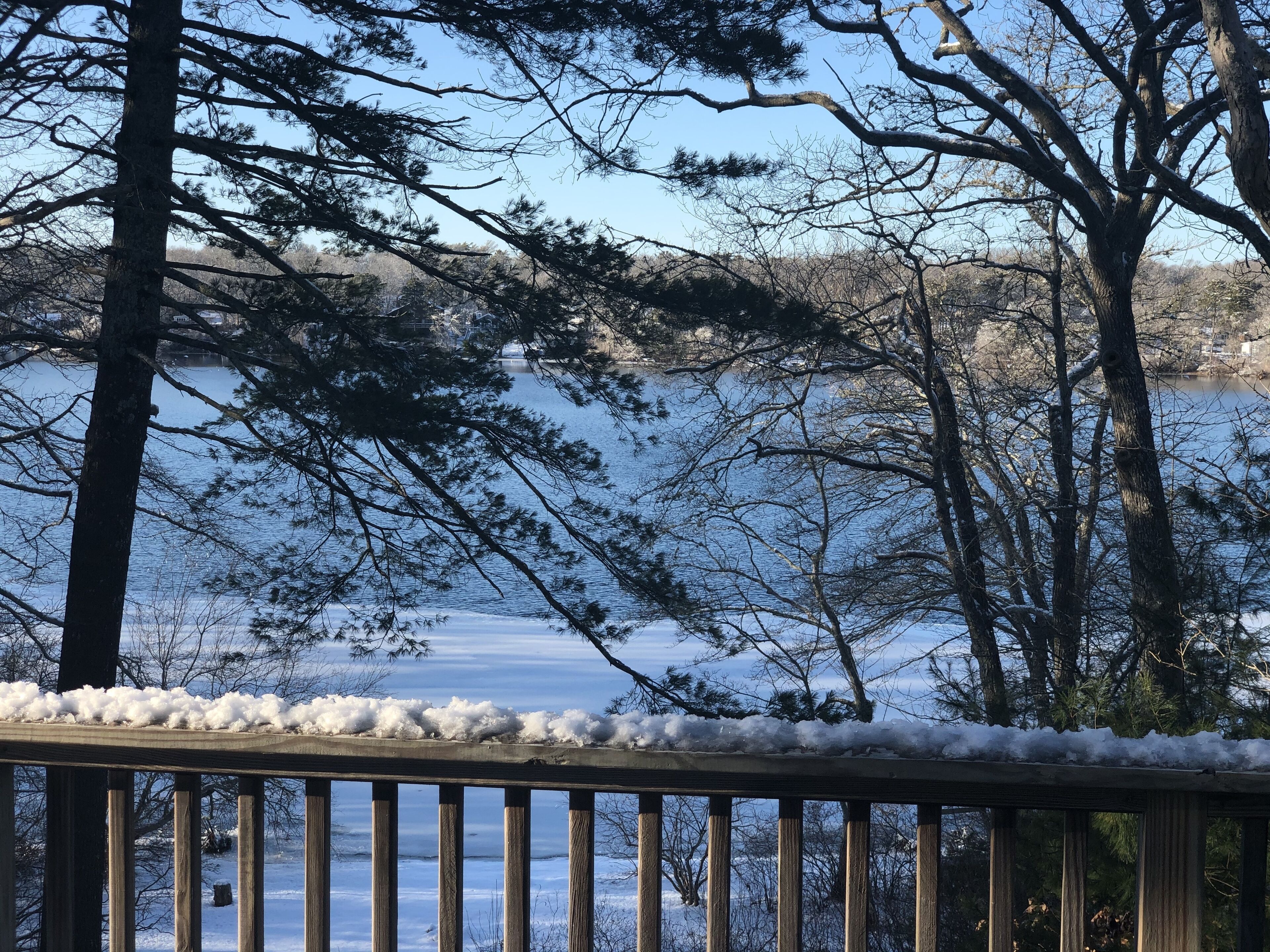 Une terrasse en bois, recouverte de neige sur la rambarde, donnant sur un lac avec des arbres en arrière-plan.
