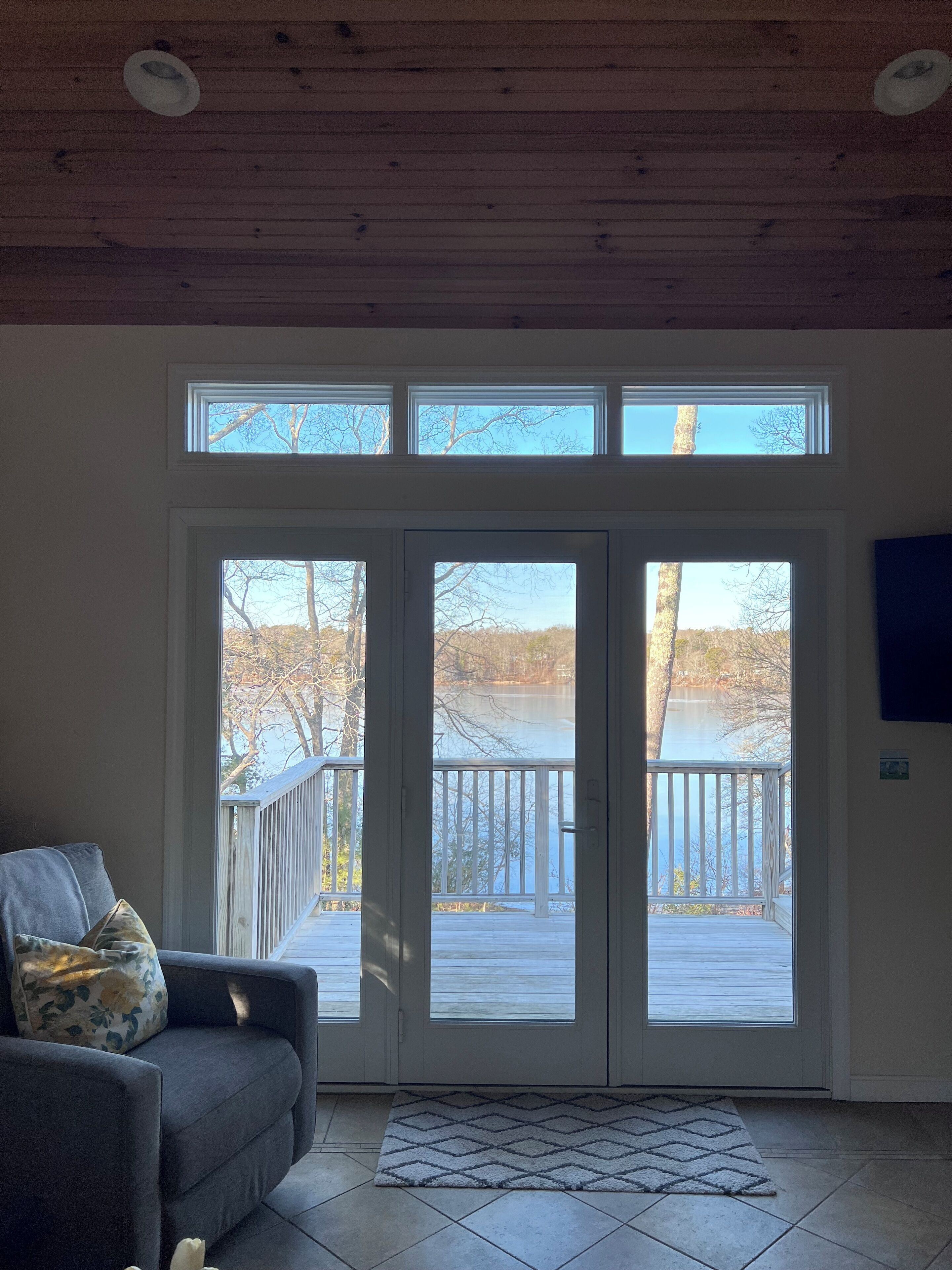 Une pièce avec un canapé gris, un tapis à motifs et une vue sur une terrasse en bois et un lac, visible à travers de grandes portes-fenêtres coulissantes.