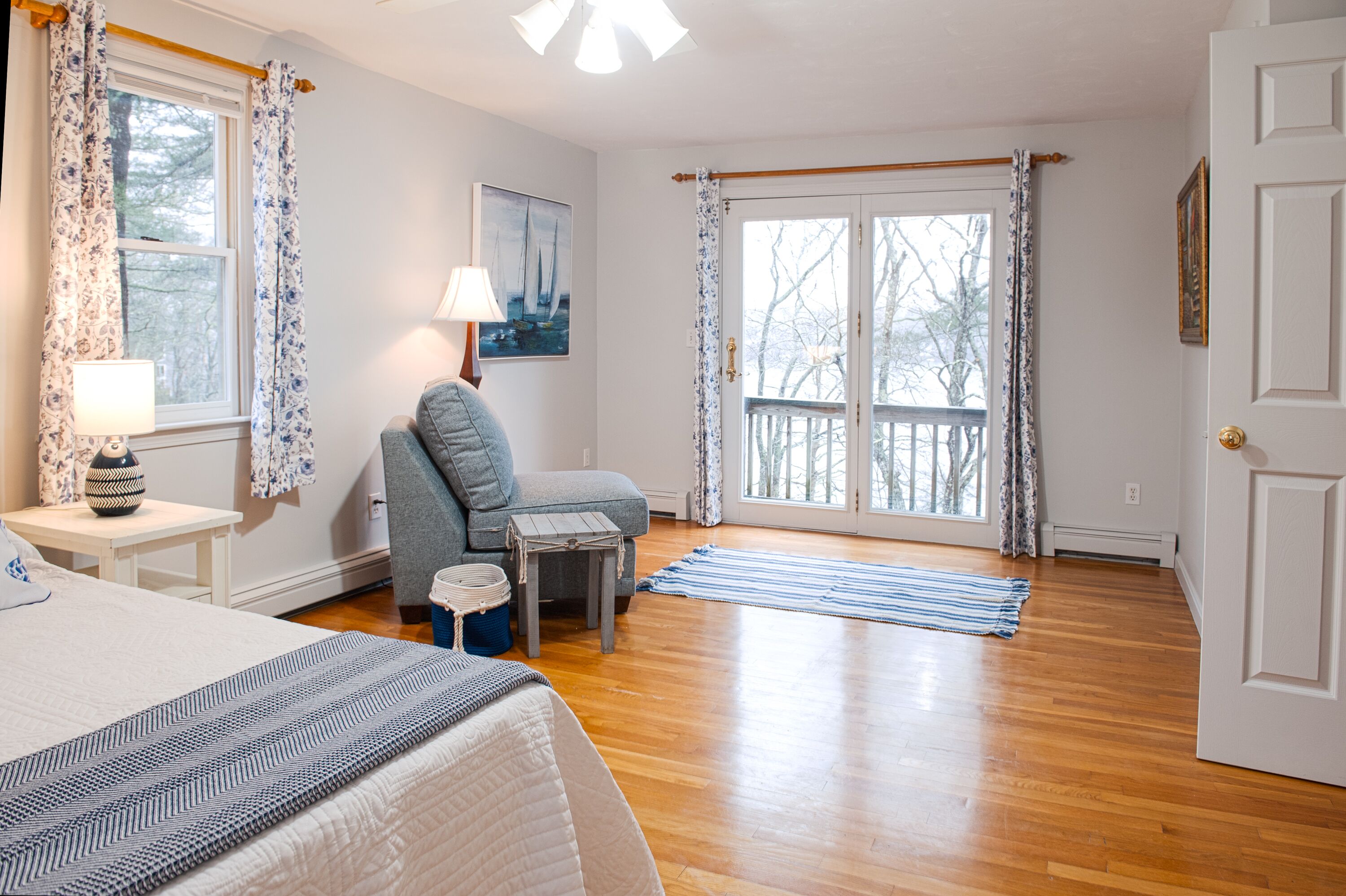 A bedroom with a bed, a gray armchair, a small table with a lamp, a window with curtains, and a door.