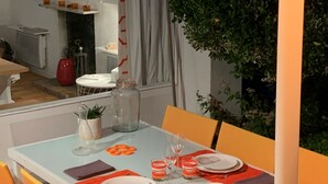 Un coin repas avec une table dressée pour quatre personnes, entourée de chaises. On aperçoit une plante en pot sur la droite et une lampe au-dessus de la table.