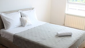 Un letto rifatto con lenzuola bianche e una coperta grigia, una finestra con vista sugli edifici e un termosifone sulla parete.