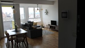Un soggiorno moderno con divano, tavolo da pranzo con sedie, televisione e vista sulla città attraverso ampie finestre.