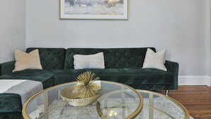 Una sala de estar con un sofá de terciopelo verde oscuro, una mesa de centro redonda de vidrio y un cuadro abstracto enmarcado en la pared.