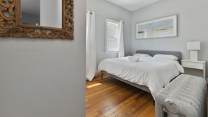 Un dormitorio con suelo de madera, una cama con ropa de cama blanca, una mesita de noche blanca, una lámpara blanca y un cuadro en la pared.