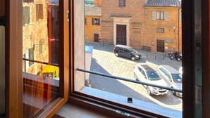A window view of a street with parked cars and a building with a brick facade. - Centralissimo Casa Piazzetta (Siena)