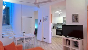 A modern living room with a large orange sofa, a dining area with a white table and chairs, a kitchen with white cabinets, and a flat-screen TV on a white stand. - Centralissimo Casa Piazzetta (Siena)