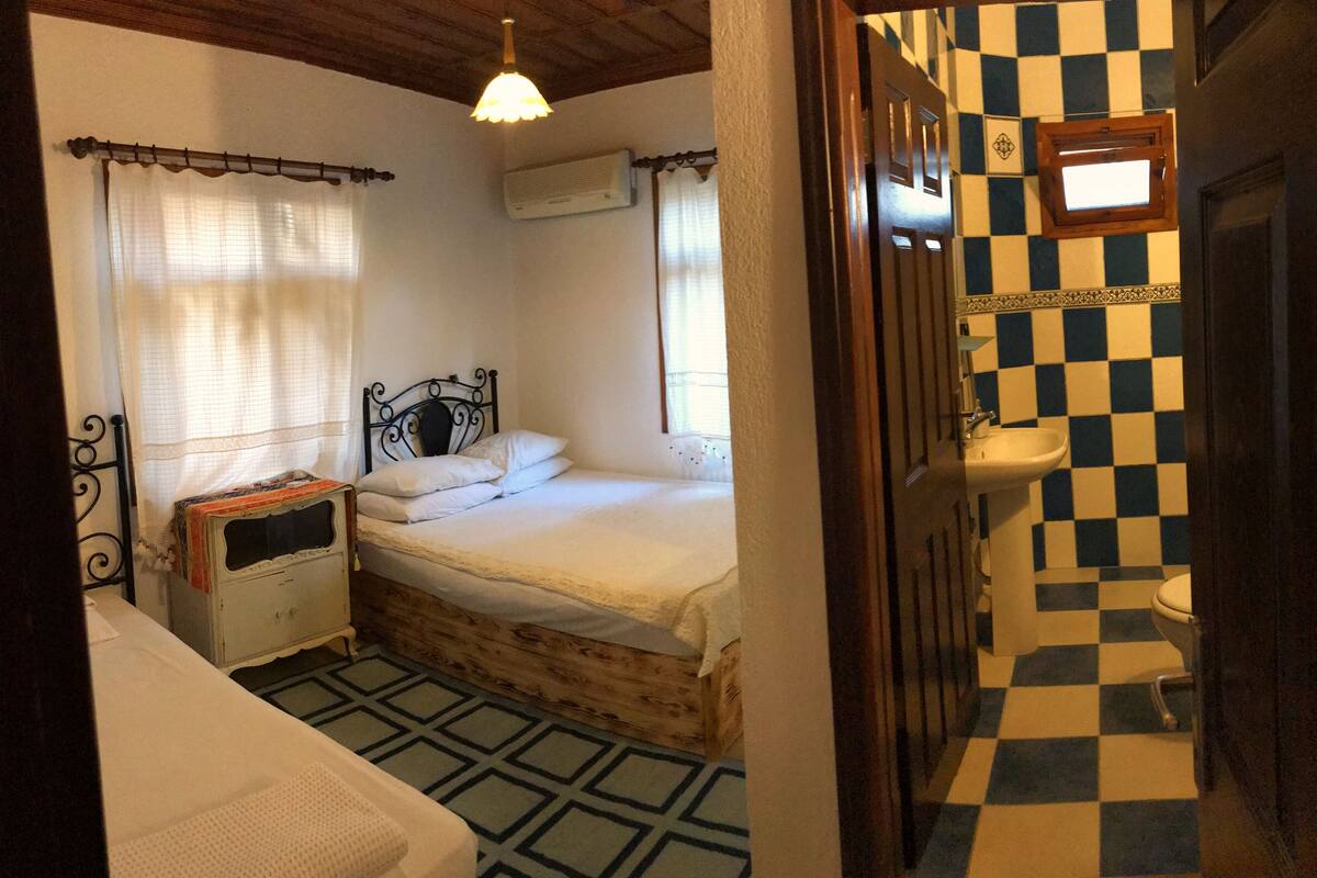 A bedroom with a wooden bed, a nightstand, a window with curtains, and a checkered bathroom floor.