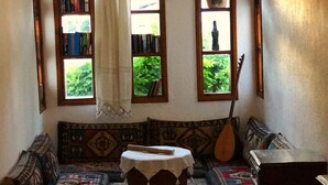 Ein gemütliches Wohnzimmer mit einer Couch, einem kleinen Tisch und einer Gitarre. Zwei Fenster mit Vorhängen und eine Bücherwand mit Büchern.