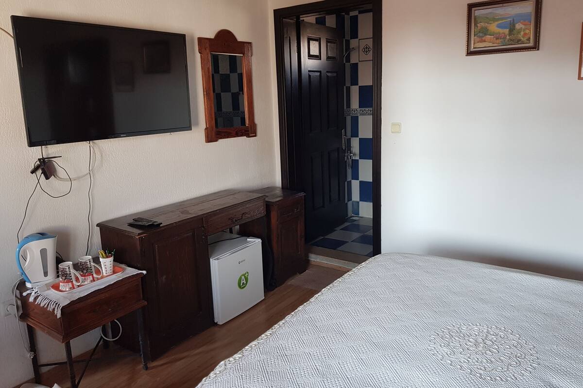 A hotel room with a bed, a TV mounted on the wall, a desk with a refrigerator, and a door leading to a bathroom with checkered tiles.